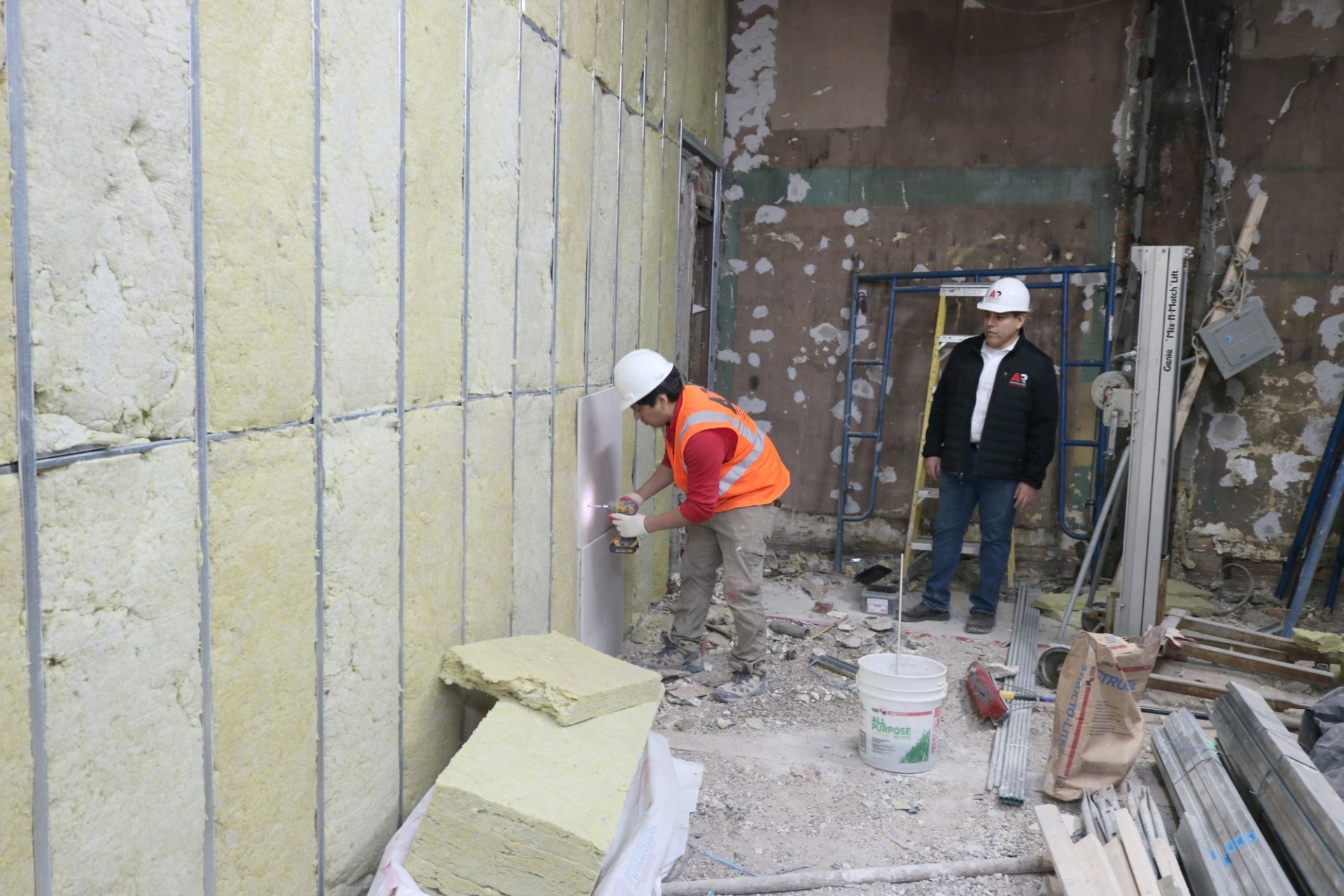 Allan Suarez standing next to a construction worker in a space being renovated, who is installing panels on a wall that is filled with insulation