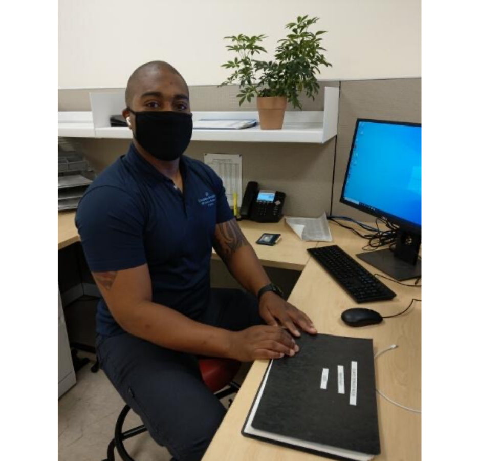 A man wearing a blue polo and mask sits in front of a desktop computer at an office cubicle