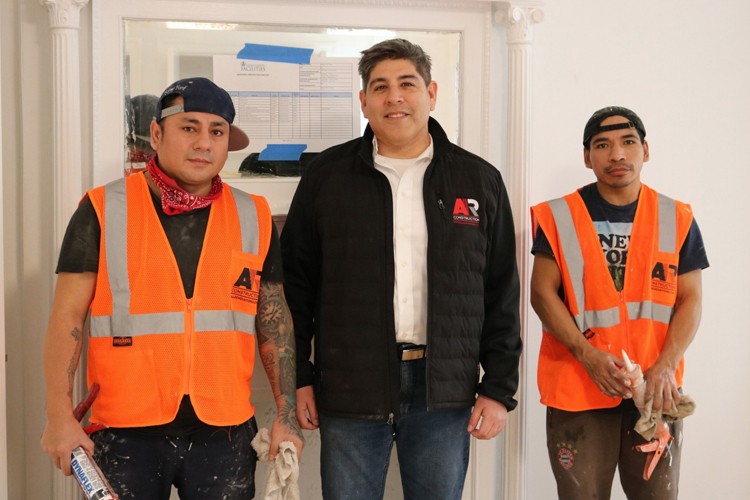 Allan Suarez standing next to two construction workers wearing hats and orange vests who are standing next to a white wall with a faux fireplace.