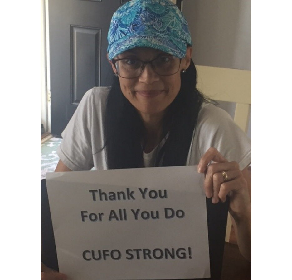 A woman wearing a blue paisley baseball cap holds a sign that says, "Thank you for all you do. CUFO strong!"