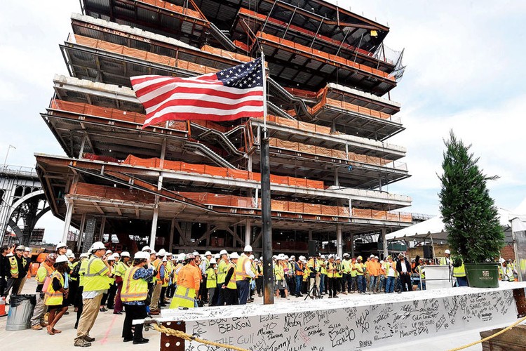 A signed steel beam with an American flag on top, with a crowd of people celebrating the topping out of the new Columbia Business School buildings, one of which is in the background
