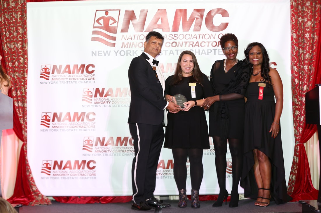 A woman accepts an award from three people who are standing in front of a white screen that has a logo that says NAMC 