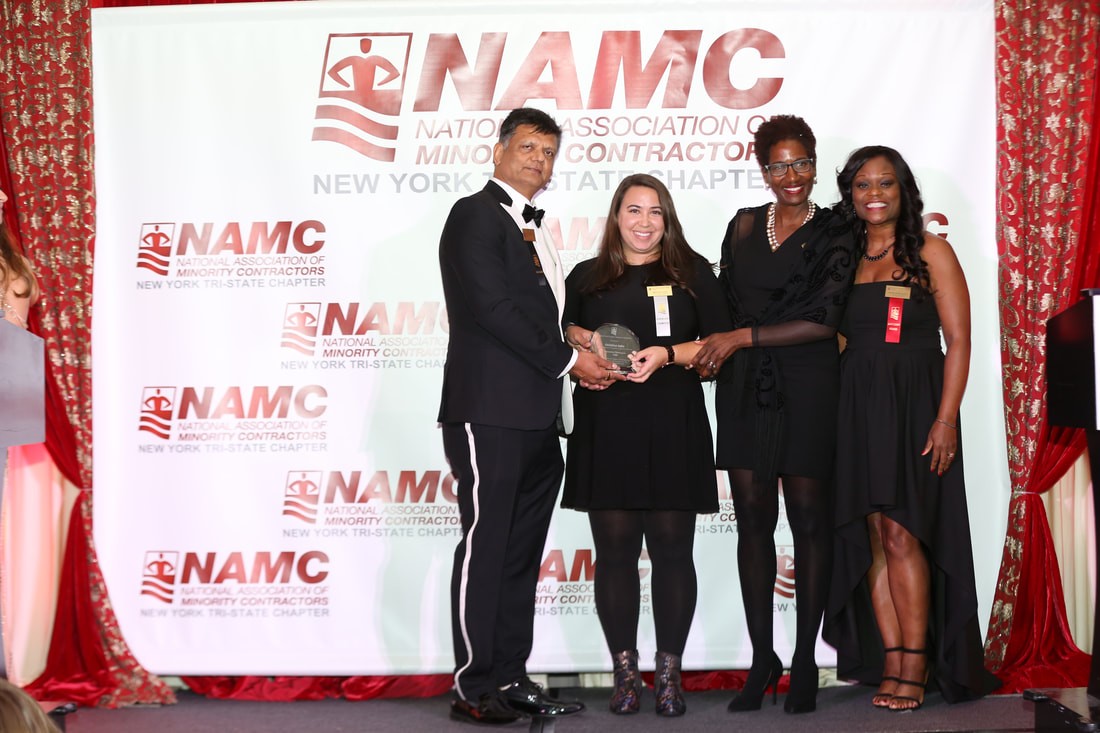 A woman accepts an award from three people who are standing in front of a white screen that has a logo that says NAMC