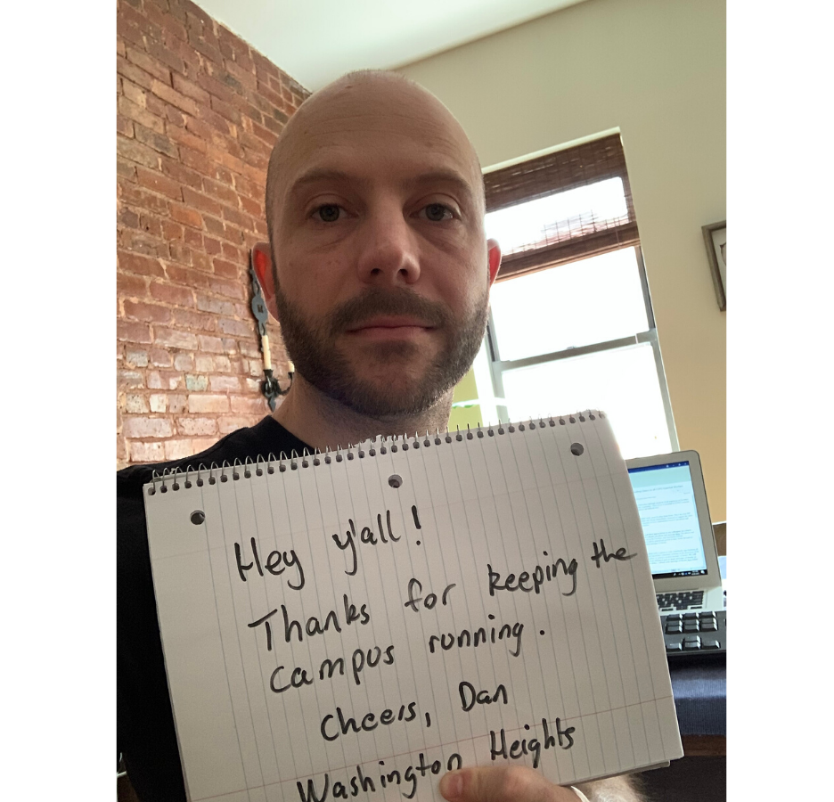 A bearded man holding a sign that reads, "Hey y'all, thanks for keeping the campus running.  Cheers, Dan - Washington Heights" in an apartment with a brick wall