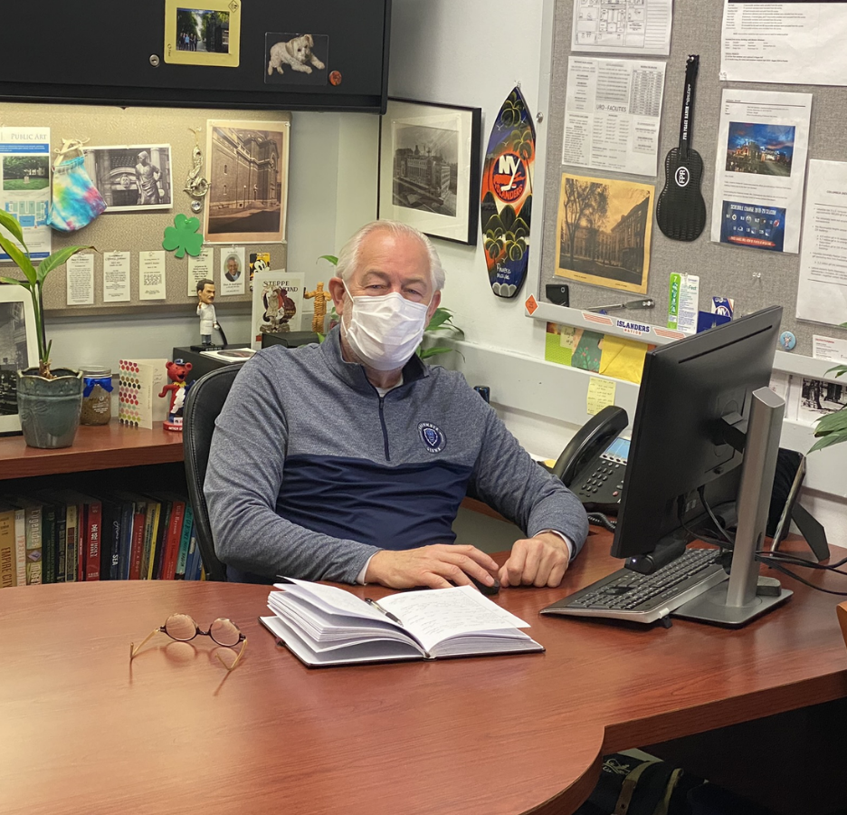 A man wearing a mask sits at a desk in an office that has memorabilia posted to the wall