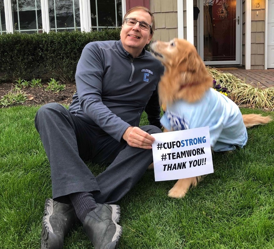 A smiling man sitting on the grass, holding a thank you sign while his golden retreiver tries to lick his face