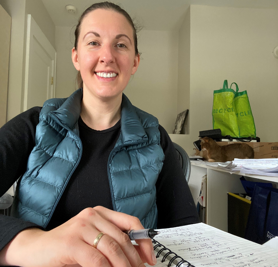 A smiling woman wearing a blue puffy vest sits in front of a notebook with a pen in her hand