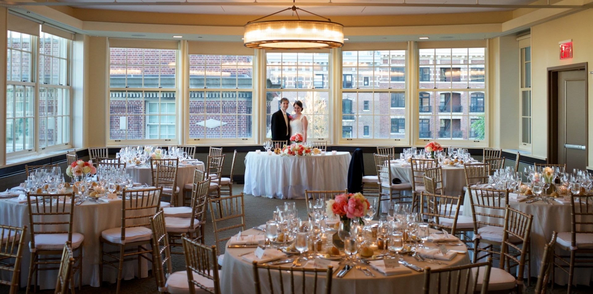 The Faculty House Presidential Ballroom set up as a wedding reception venue, with table set up throughout the room and the bride and groom standing in the back, smiling.