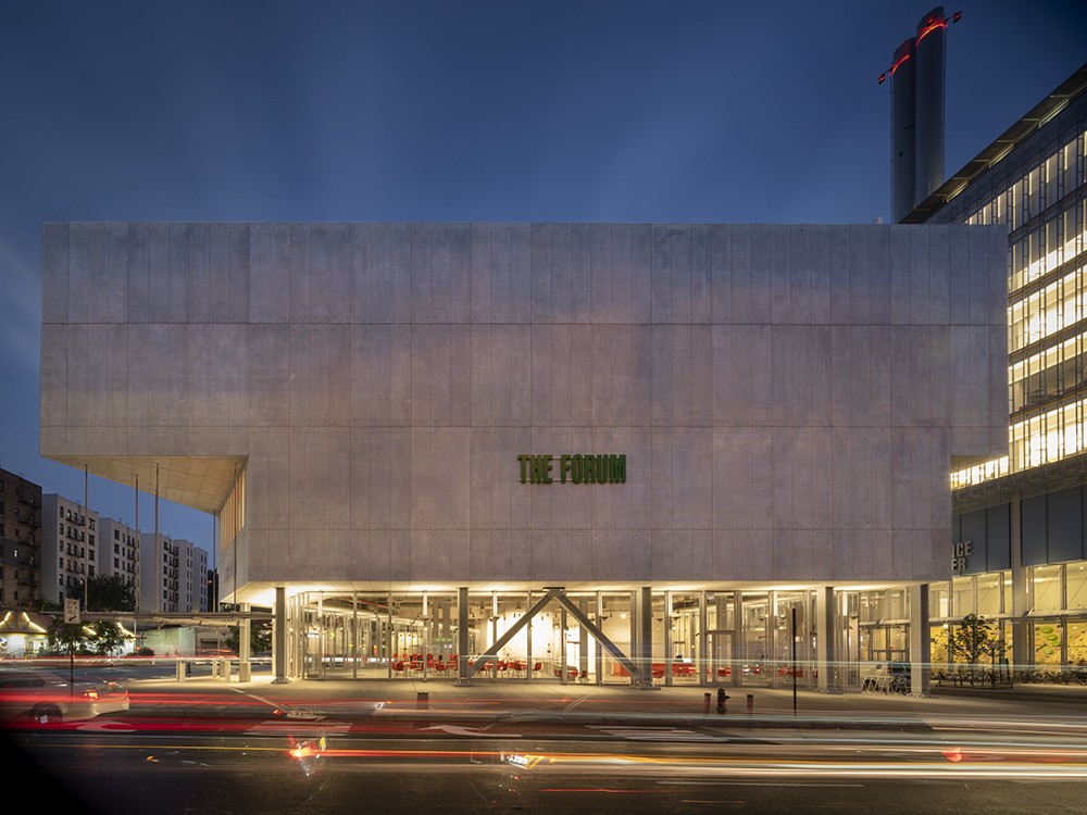 Photo: Nic Lehoux The side of The Forum building in the evening, with lights illuminated on the ground floor and a green sign that reads, "The Forum" in the middle of the facade.