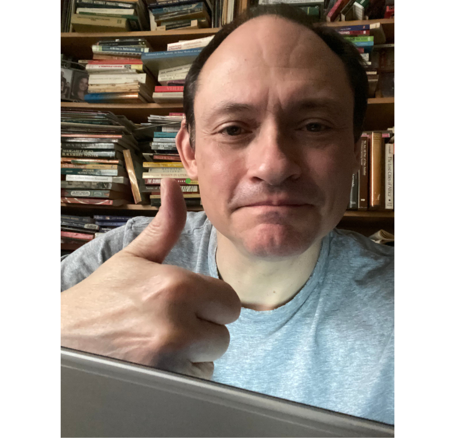 A man looks into the camera and gives a thumbs up in front of a wall full of books