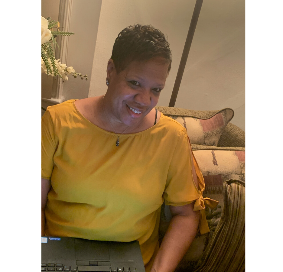A woman wearing a yellow shirt with a laptop on her lap smiles at the camera