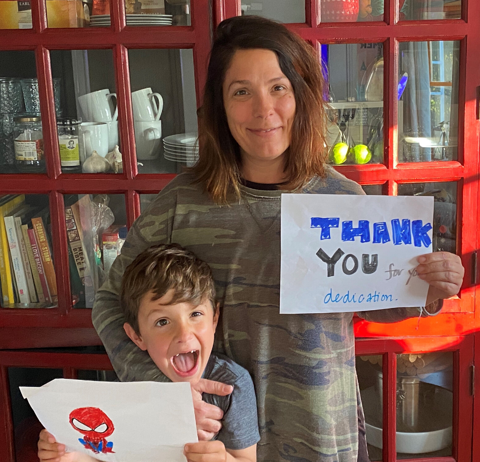 A young boy holding a handmade picture of Spiderman stands next to his mother who is holding a handmade thank you sign.