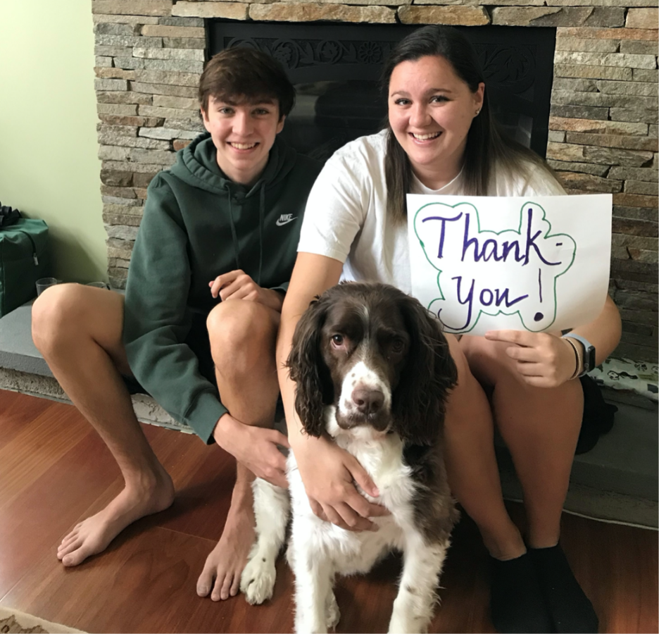 Two teenagers sit with their white and brown dog in front of a fireplace with a Thank You sign
