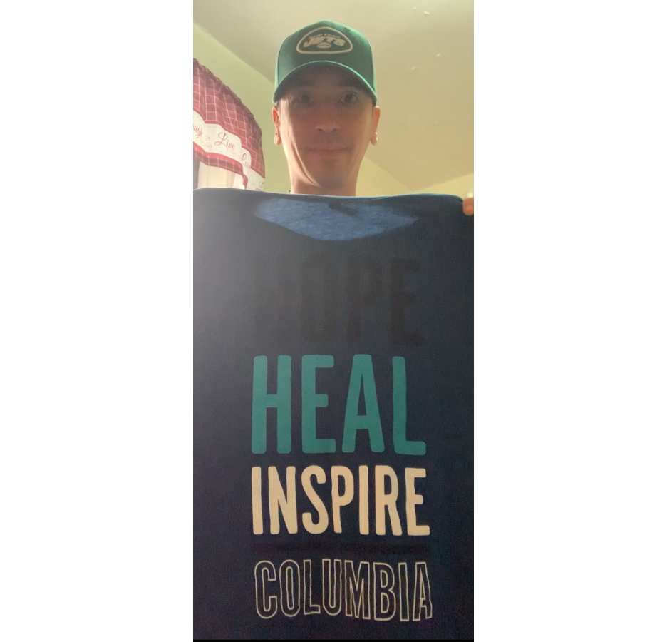 A man wearing a green Jets baseball cap holds a shirt that says, "Hope, heal, inspire, Columbia"