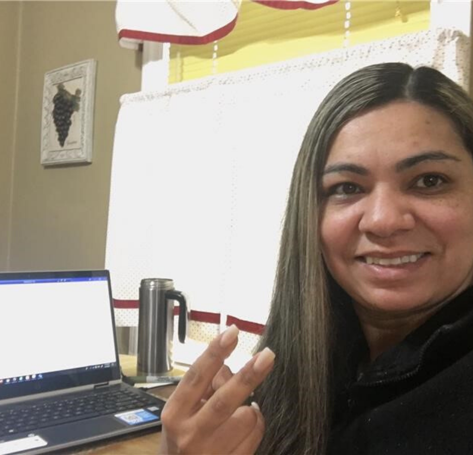 A woman with brown hair makes a peace sign. She sits in front of a laptop.