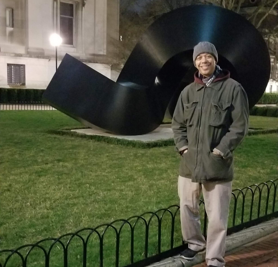 A person wearing a green jacket stands in front of an abstract sculpture on front of Uris Lawn.