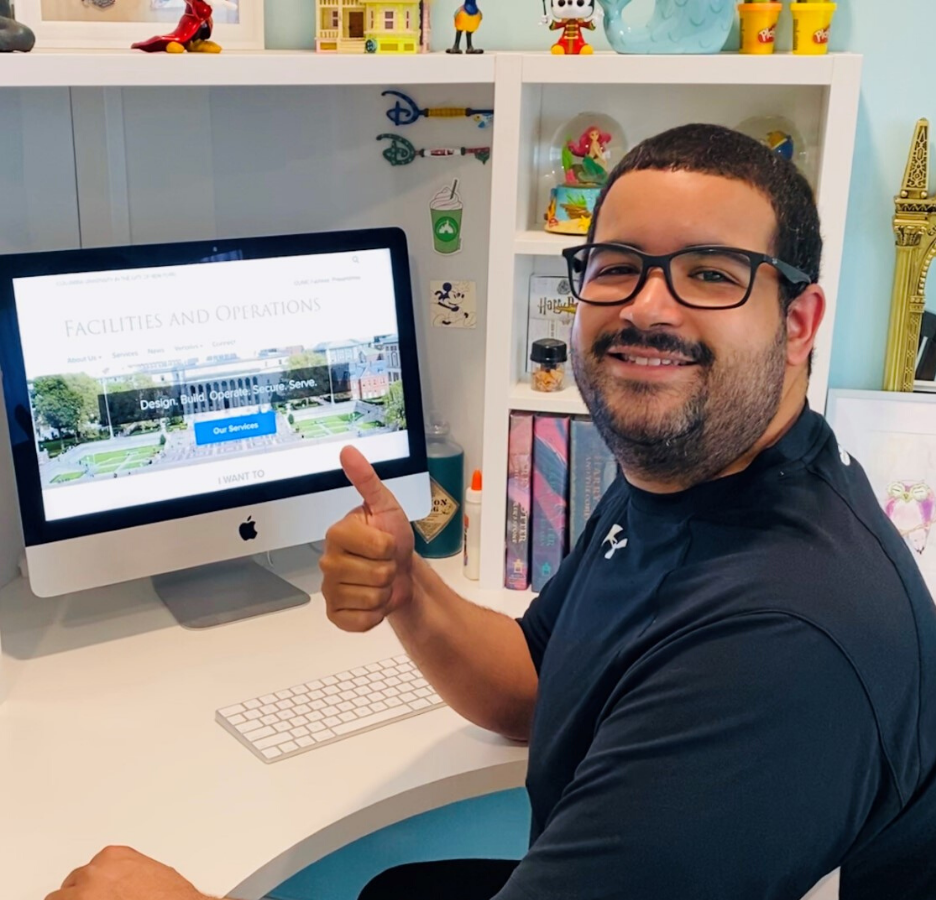 A person wearing glasses and a dark blue t-shirt makes a thumb up sign. He sits at a desk. A computer monitor is behind him.