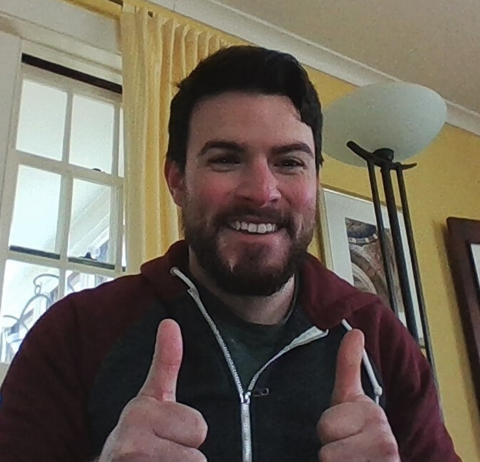 A person with dark hair and a beard wearing a maroon hoodie, smiling and giving two thumbs up 