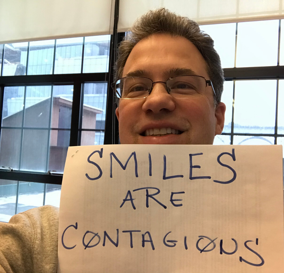 A man with glasses smiles and holds up sign that says "Smiles are contagious"