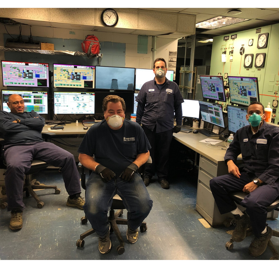 A group of people with masks on sitting in a computer room with multiple computer monitors with detailed information displayed on the desk and multiple clocks on the wall.