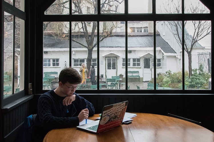 A student sitting at a table looking at his laptop in a windowed room with a courtyard and white building in the background