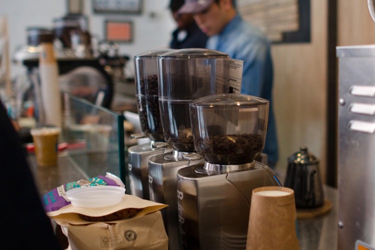 The interior of a cafe with cups and coffee grinders in the forefront and two workers preparing drinks next to equipment in the background