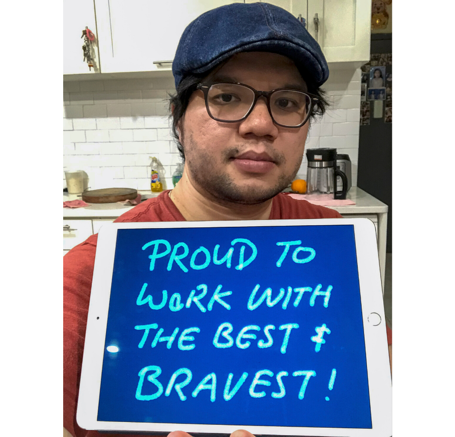 A person wearing a blue hat and glasses holds up a sign that says, "proud to work with the bravest."
