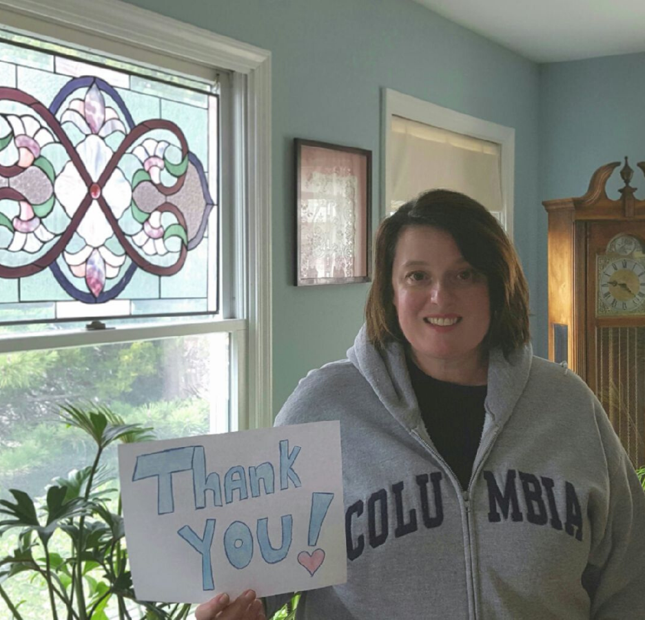 A woman wearing a grey Columbia-branded jacket holds a handmade thank you sign in her home next to a stained glass window, plant, and grandfather clock