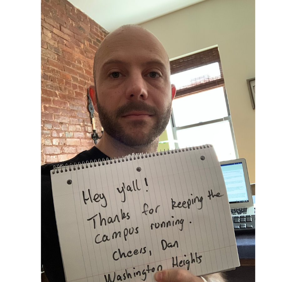 A bearded man holding a sign that reads, "Hey y'all, thanks for keeping the campus running. Cheers, Dan - Washington Heights" in an apartment with a brick wall