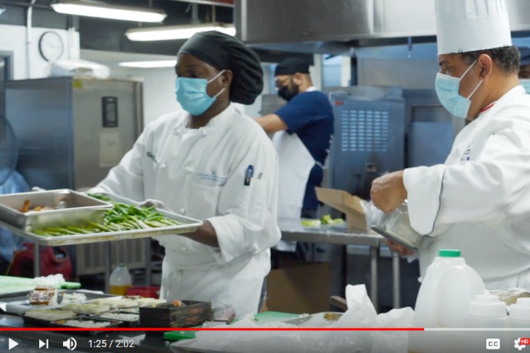 Columbia Dining employees work to prepare food in a kitchen, with two people in the forefront holding trays of prepped food