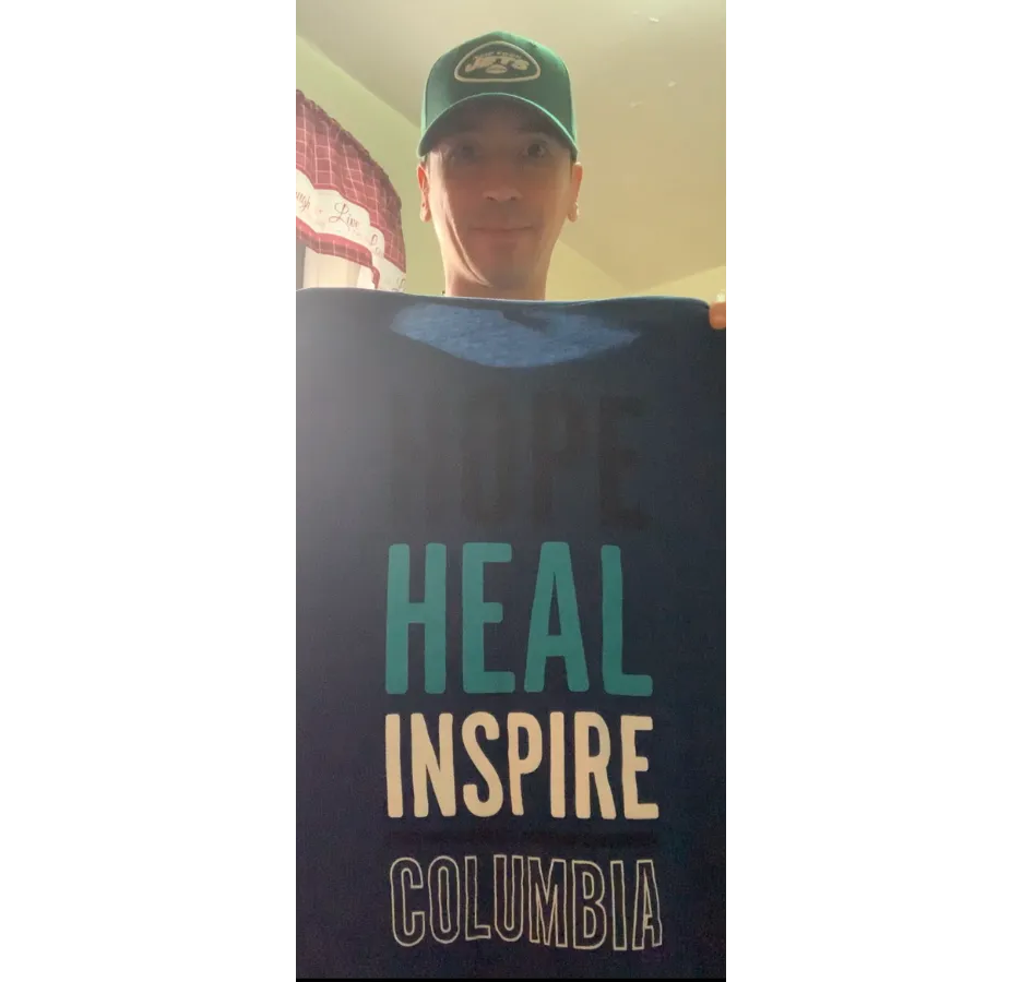 A man wearing a green Jets baseball cap holds a shirt that says, "Hope, heal, inspire, Columbia"