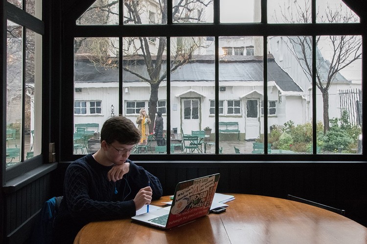 A student sitting at a table looking at his laptop in a windowed room with a courtyard and white building in the background