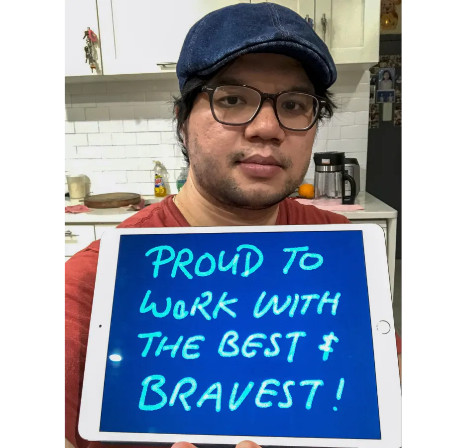 A person wearing a blue hat and glasses holds up a sign that says, "proud to work with the bravest."